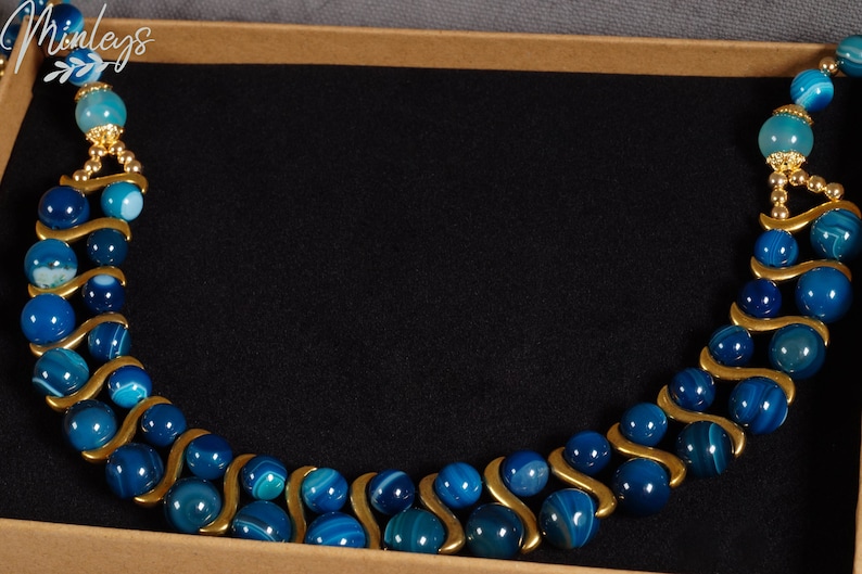 May include: A blue and gold agate beaded necklace with a wavy design. The necklace is displayed in a brown box.