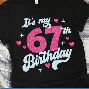 Op de afbeelding: Zwart T-shirt met de tekst "It's my 67th Birthday" in roze en blauwe glitterletters. Het shirt is versierd met kleine roze hartjes en sterren.