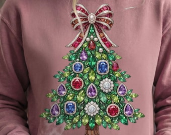 May include: Dusty rose sweatshirt featuring a Christmas tree design. The tree is adorned with colorful gemstones and a large bow at the top. The tree trunk is brown, and the gemstones are red, blue, purple, and white.