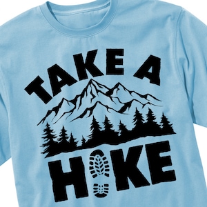 Op de afbeelding: Een lichtblauw t-shirt met een zwart grafisch ontwerp met de tekst "Take a Hike" met een bergketen en een voetafdruk.