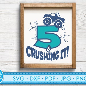 May include: A framed print with a white background features a teal number 5 with a monster truck on top. The phrase "& CRUSHING IT!" is below the number in navy blue. The frame is made of wood.