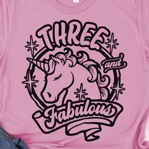 May include: Pink t-shirt with a black graphic design of a unicorn with a horn, stars, and the text "Three and Fabulous".
