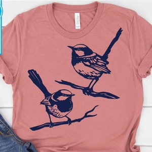 Op de afbeelding: Een roze T-shirt met een ontwerp van twee blauwe vogels die op takken zitten.