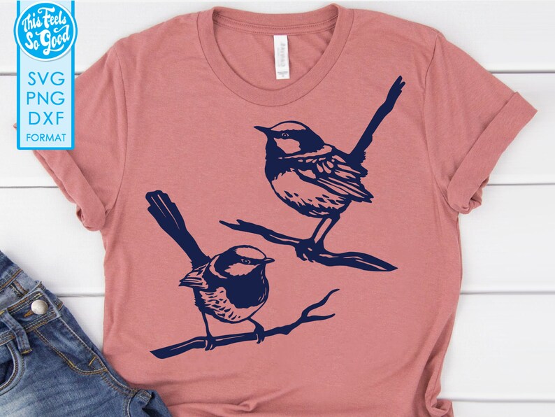 Op de afbeelding: Een roze T-shirt met een ontwerp van twee blauwe vogels die op takken zitten.