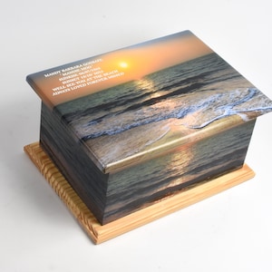 May include: A wooden box with a printed image of a beach sunset. The box has a light-colored wooden base. Text on the box reads "MANDY BARBARA GODSARY, MANDIE MOO, SUNRISE 06/07/15, SUNSET 19/10/15, WELL SEE YOU AT THE BEACH, ALWAYS LOVED FOREVER MISSED."