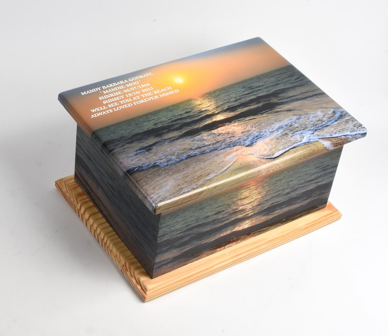 May include: A wooden box with a printed image of a beach sunset. The box has a light-colored wooden base. Text on the box reads "MANDY BARBARA GODSARY, MANDIE MOO, SUNRISE 06/07/15, SUNSET 19/10/15, WELL SEE YOU AT THE BEACH, ALWAYS LOVED FOREVER MISSED."