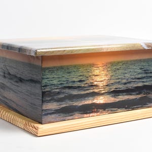 May include: A rectangular wooden box with a sunset ocean scene printed on its sides and top. The box sits on a light-colored wooden base. The sunset features a golden glow reflecting on the water.