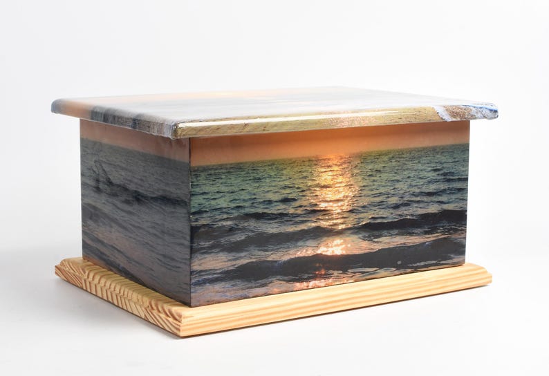 May include: A rectangular wooden box with a sunset ocean scene printed on its sides and top. The box sits on a light-colored wooden base. The sunset features a golden glow reflecting on the water.