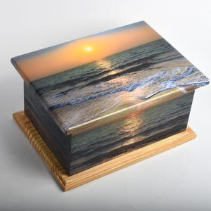 May include: A rectangular wooden box with a lid, featuring a scenic image of a sunset over the ocean. The box has a light-colored wooden base and a glossy finish, with the sunset image wrapping around the sides.