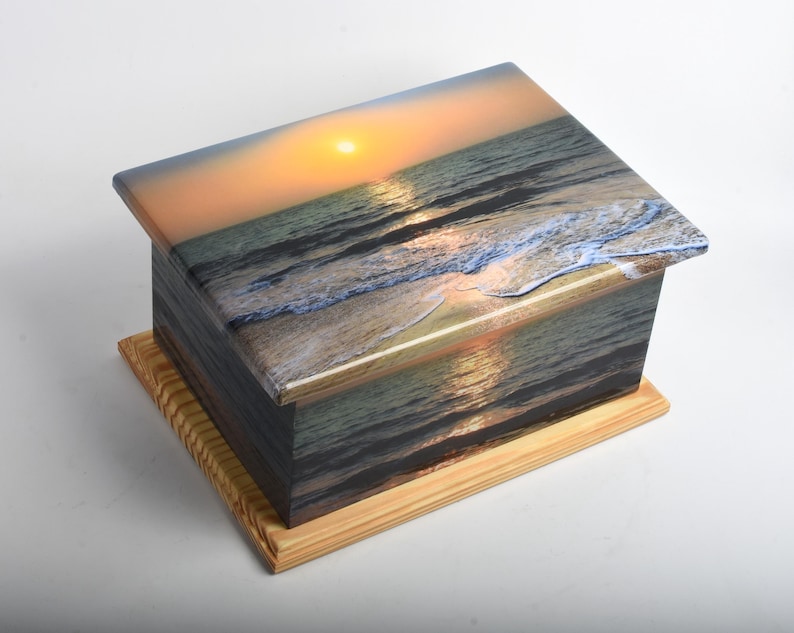 May include: A rectangular wooden box with a lid, featuring a scenic image of a sunset over the ocean. The box has a light-colored wooden base and a glossy finish, with the sunset image wrapping around the sides.