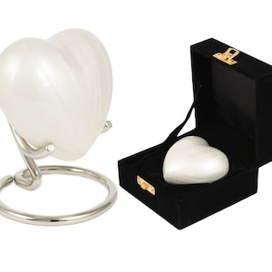 May include: A pearlescent white heart-shaped urn, presented on a silver metal stand, and another inside a black velvet box with a gold clasp. The urn has a smooth, glossy finish and a classic heart shape.