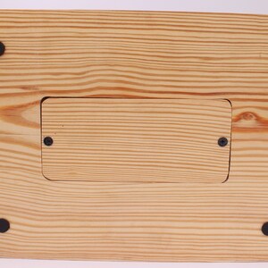 May include: A wooden box with a natural finish, featuring visible wood grain and a rectangular recessed area secured with two screws. Four black rubber feet are attached to the base for stability.