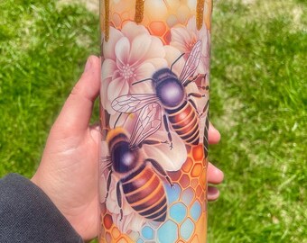 May include: A stainless steel tumbler with a gold glitter finish and a design of bees and honeycomb. The design features two bees with black and brown stripes, and a honeycomb pattern with white and blue accents. The tumbler is decorated with a dripping gold glitter effect.