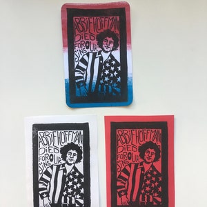 Puede incluir: Tres impresiones de linóleo en blanco y negro de un retrato de Abbie Hoffman con el texto "Abbie Hoffman Died For Our Sins". Las impresiones están en papel blanco, rojo y azul.