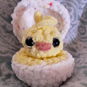 May include: A yellow chick plush toy with black eyes and a pink beak, sitting inside a white crocheted egg with colorful sprinkles.