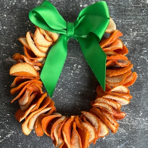 May include: A circular wreath made of dried orange slices, with a bright green satin bow at the top. The orange slices are arranged in a ring shape, and the bow adds a pop of color against the natural tones of the wreath.