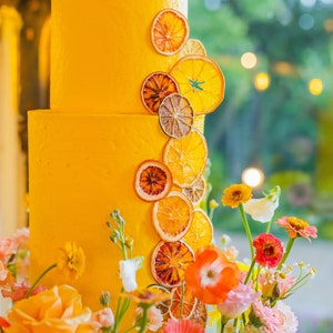 K&ouml;nnte beinhalten: Eine gelbe Hochzeitstorte, verziert mit getrockneten Orangenscheiben und Blumen. Die Torte steht auf einem durchsichtigen St&auml;nder und ist von Blumen in Glasvasen umgeben.