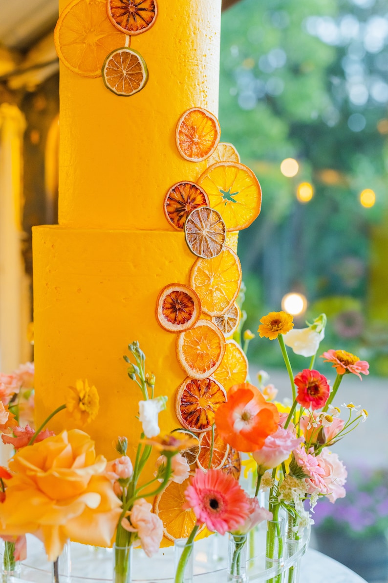 K&ouml;nnte beinhalten: Eine gelbe Hochzeitstorte, verziert mit getrockneten Orangenscheiben und Blumen. Die Torte steht auf einem durchsichtigen St&auml;nder und ist von Blumen in Glasvasen umgeben.