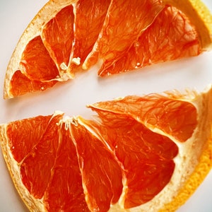 May include: Two dried grapefruit slices, showcasing vibrant orange-red segments and a light-colored rind. The fruit's texture is visible, with a focus on the natural patterns and details. The slices are arranged on a white surface.