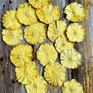 May include: A collection of dried pineapple slices arranged on a rustic wooden surface. The slices are a vibrant yellow color and have a natural, sun-dried appearance.