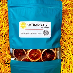 K&ouml;nnte beinhalten: Ein blauer wiederverschlie&szlig;barer Beutel mit einem Sichtfenster, das getrocknete Orangenscheiben zeigt. Der Beutel hat ein wei&szlig;es Etikett mit dem Text "KATRAM COVE pretty little things!" und einem QR-Code. Der Text "Store airtight up to 1 year, scan to reorder" befindet sich ebenfalls auf dem Etikett.