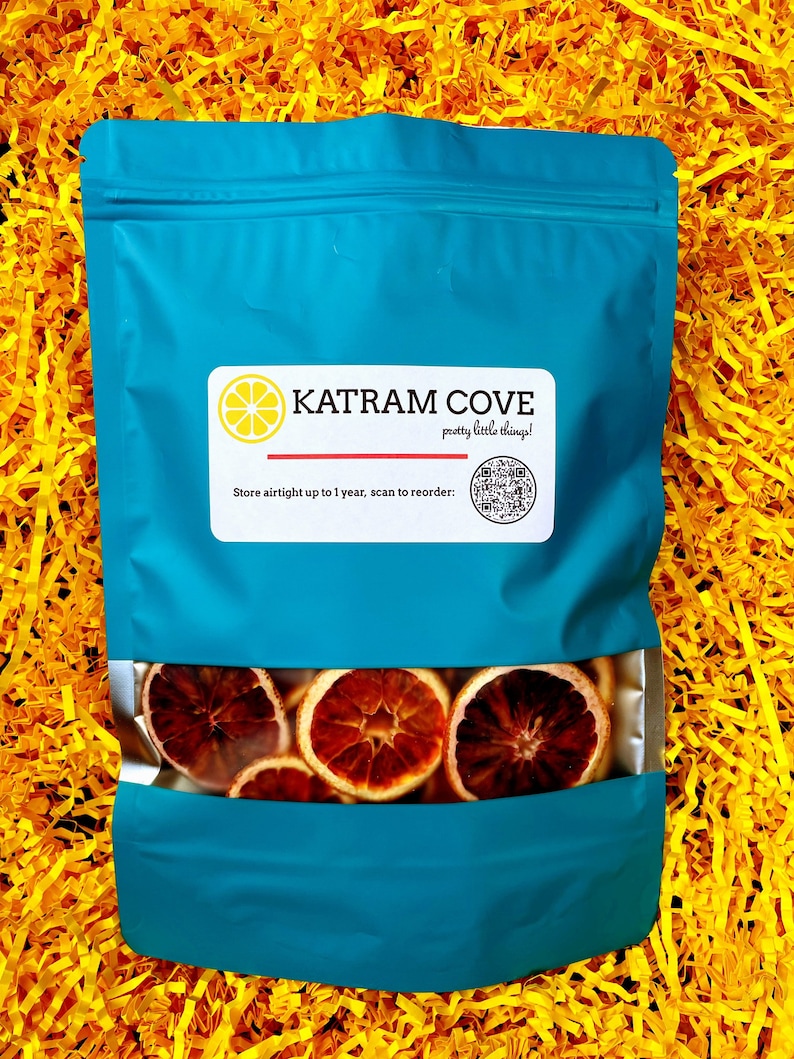 K&ouml;nnte beinhalten: Ein blauer wiederverschlie&szlig;barer Beutel mit einem Sichtfenster, das getrocknete Orangenscheiben zeigt. Der Beutel hat ein wei&szlig;es Etikett mit dem Text "KATRAM COVE pretty little things!" und einem QR-Code. Der Text "Store airtight up to 1 year, scan to reorder" befindet sich ebenfalls auf dem Etikett.