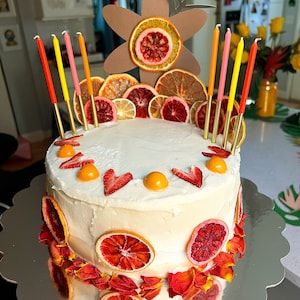 K&ouml;nnte beinhalten: Ein wei&szlig;er Kuchen, der mit getrockneten Orangenscheiben, roten Rosenbl&auml;ttern und Erdbeeren verziert ist. Der Kuchen ist mit einem bl&uuml;tenf&ouml;rmigen Pappkarton und Kerzen verziert.