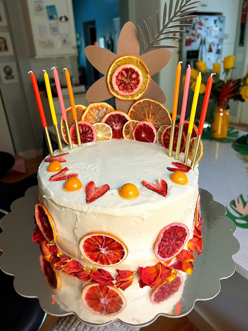 K&ouml;nnte beinhalten: Ein wei&szlig;er Kuchen, der mit getrockneten Orangenscheiben, roten Rosenbl&auml;ttern und Erdbeeren verziert ist. Der Kuchen ist mit einem bl&uuml;tenf&ouml;rmigen Pappkarton und Kerzen verziert.