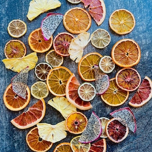May include: Overhead shot of a collection of dried fruit slices, including oranges, lemons, grapefruits, dragon fruit, and pineapple. The slices are arranged on a dark blue surface, showcasing their natural colors and textures. A vibrant and colorful display.