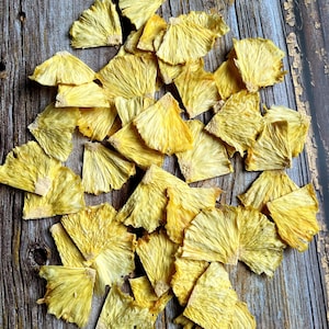 May include: A pile of dried pineapple slices on a rustic wooden surface. The pineapple slices are a light yellow color and have a slightly translucent appearance.