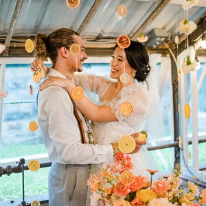 K&ouml;nnte beinhalten: Eine Braut und ein Br&auml;utigam umarmen sich auf ihrer Hochzeitsfeier. Die Braut tr&auml;gt ein wei&szlig;es Kleid mit Blumenmuster und einen Schleier. Der Br&auml;utigam tr&auml;gt ein wei&szlig;es Hemd, graue Hosen und einen braunen G&uuml;rtel. Das Paar ist umgeben von einem Blumenschmuck mit orangefarbenen und gelben Blumen. Getrocknete Zitrusscheiben h&auml;ngen als Dekoration von der Decke.