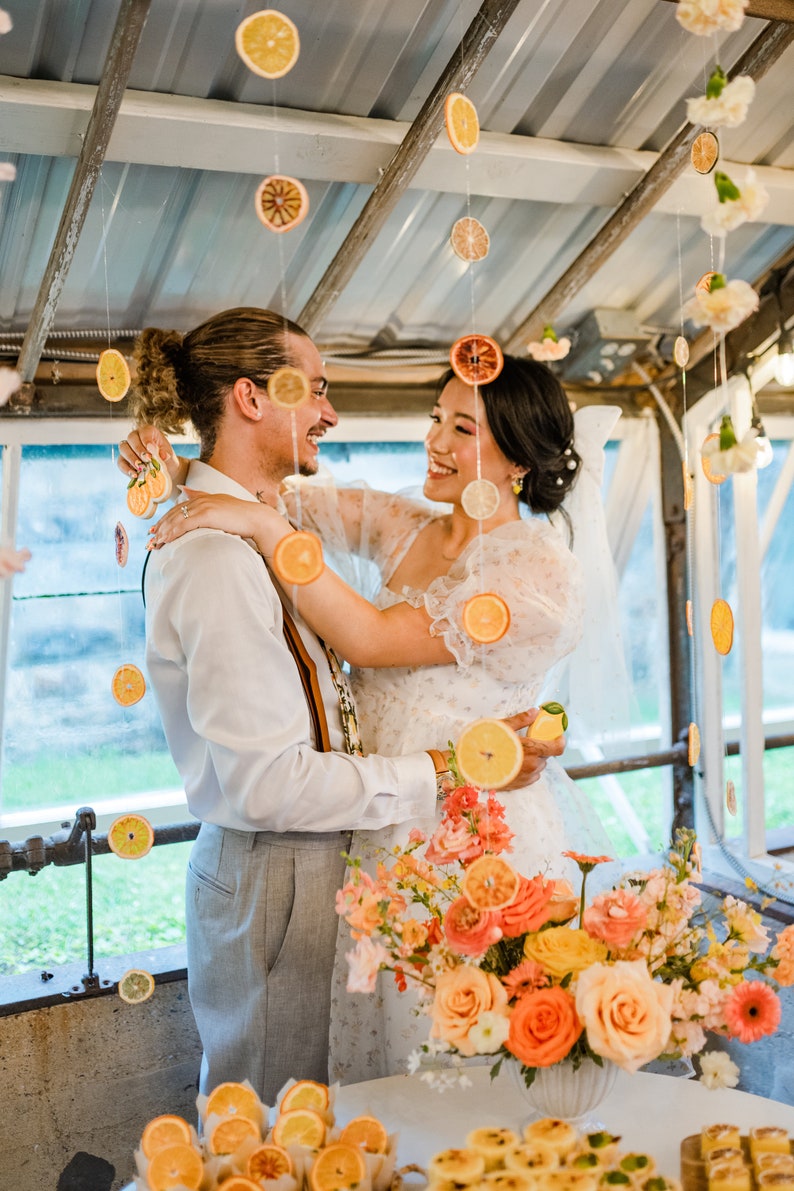 K&ouml;nnte beinhalten: Eine Braut und ein Br&auml;utigam umarmen sich auf ihrer Hochzeitsfeier. Die Braut tr&auml;gt ein wei&szlig;es Kleid mit Blumenmuster und einen Schleier. Der Br&auml;utigam tr&auml;gt ein wei&szlig;es Hemd, graue Hosen und einen braunen G&uuml;rtel. Das Paar ist umgeben von einem Blumenschmuck mit orangefarbenen und gelben Blumen. Getrocknete Zitrusscheiben h&auml;ngen als Dekoration von der Decke.