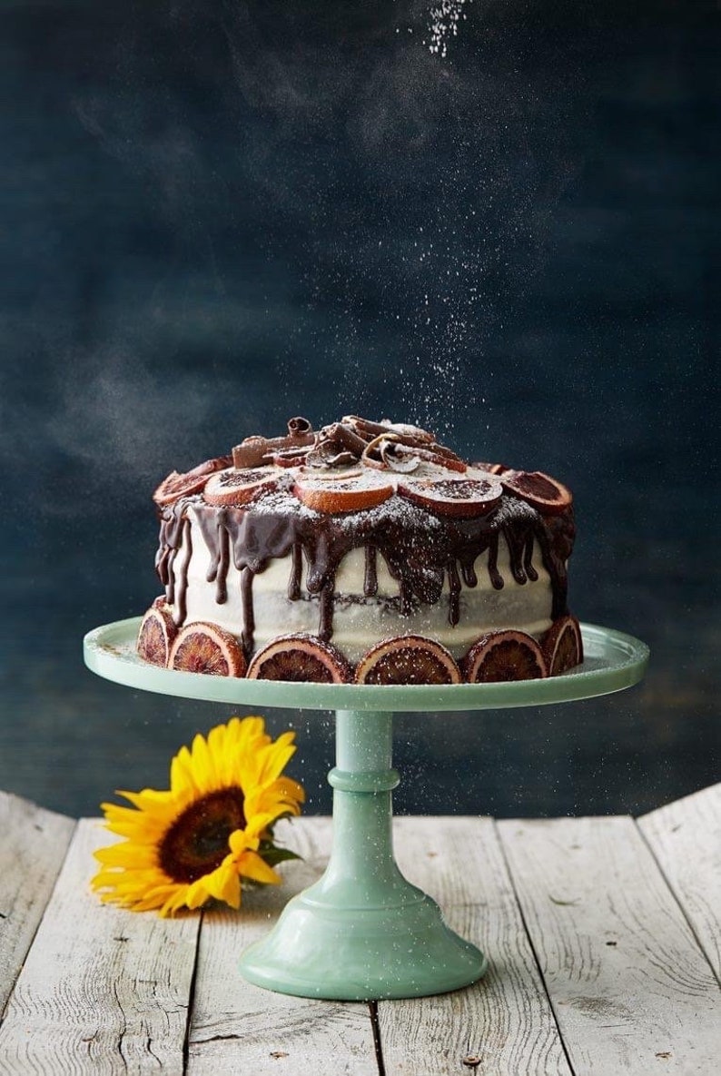 K&ouml;nnte beinhalten: Ein Schokoladenkuchen mit Orangenscheiben und Schokoladen&uuml;berzug auf einem hellblauen Kuchenst&auml;nder. Der Kuchen ist mit Puderzucker best&auml;ubt und im Vordergrund befindet sich eine Sonnenblume.