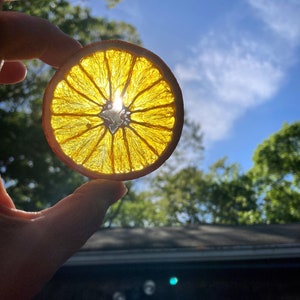 K&ouml;nnte beinhalten: Eine getrocknete Orangenscheibe, die gegen die Sonne gehalten wird, erzeugt einen leuchtend gelben Heiligenschein um die Frucht. Die Sonne scheint durch die Orangenscheibe und wirft ein helles Licht.