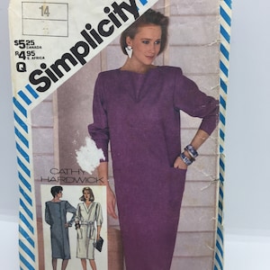 Può includere: Modello di cucito Simplicity 6105 vintage con un design di abito viola di Cathy Hardwick. La busta del modello mostra una donna che indossa l'abito, con una taglia 14 indicata. La busta ha un bordo a righe blu e bianche.