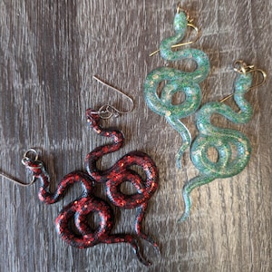 May include: A pair of red glitter snake earrings and a pair of green glitter snake earrings. The earrings are made of resin and have a dangling design.