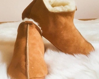 Puede incluir: Un par de zapatillas de ante marrón con forro de piel de oveja blanca. Las zapatillas están sobre una superficie blanca y esponjosa.