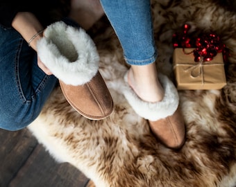 May include: A pair of brown suede slippers with white faux fur lining. The slippers are being worn by a person sitting on a faux fur rug.