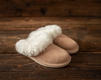 May include: A pair of beige suede slippers with white faux fur trim. The slippers are on a wooden surface.