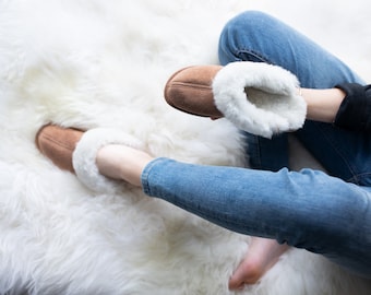 May include: A pair of brown suede slippers with white faux fur lining. The slippers are being worn by a person sitting on a white fluffy rug.