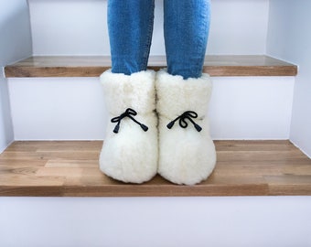 May include: A pair of white sheepskin slippers with black ribbon ties. The slippers are on a wooden step.