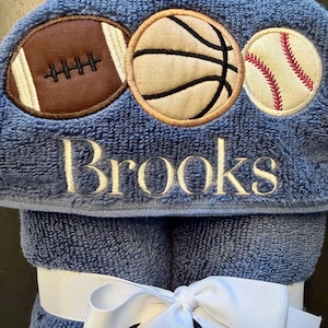 May include: A blue hooded towel featuring embroidered appliques of a brown football, a tan basketball, and a white baseball. The name Brooks is embroidered in white thread below the sports balls. The towel is tied with a white ribbon bow.