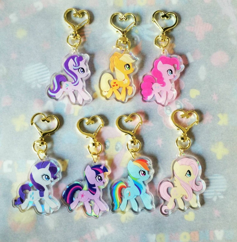 May include: Set of 8 colorful My Little Pony keychains featuring different characters. Each keychain has a gold heart-shaped ring and a clear plastic charm with a detailed image of a pony. The ponies are pink, purple, blue, yellow, orange, and white.
