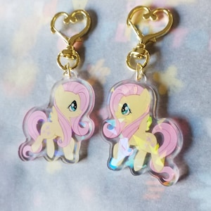 May include: Two pink and yellow cartoon pony charms with gold heart-shaped keychains. The charms are iridescent and feature a pink pony with a yellow mane and tail.