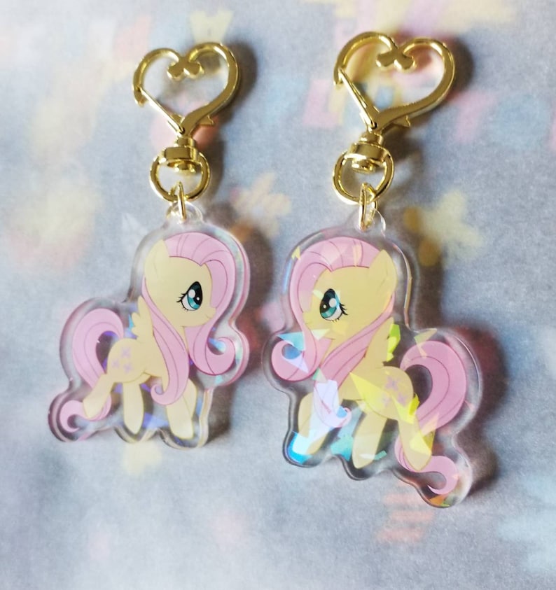 May include: Two pink and yellow cartoon pony charms with gold heart-shaped keychains. The charms are iridescent and feature a pink pony with a yellow mane and tail.
