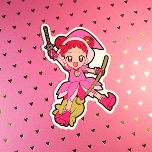 May include: A pink sticker featuring a cartoon witch character with red hair, wearing a pink dress and hat, holding a sword and a broom. The background is pink with gold heart shapes.