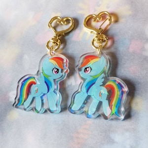 May include: Two gold keychains with heart-shaped rings feature clear acrylic charms of Rainbow Dash from My Little Pony. The charms show Rainbow Dash in a light blue pose with a rainbow tail and wings.