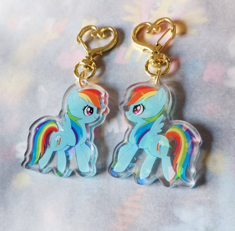 May include: Two gold keychains with heart-shaped rings feature clear acrylic charms of Rainbow Dash from My Little Pony. The charms show Rainbow Dash in a light blue pose with a rainbow tail and wings.