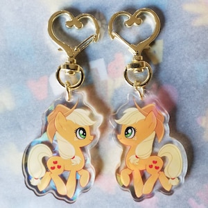 May include: Two gold heart-shaped keychains with clear acrylic charms featuring a cartoon image of Applejack from My Little Pony. The charms are translucent and show a glittery effect.