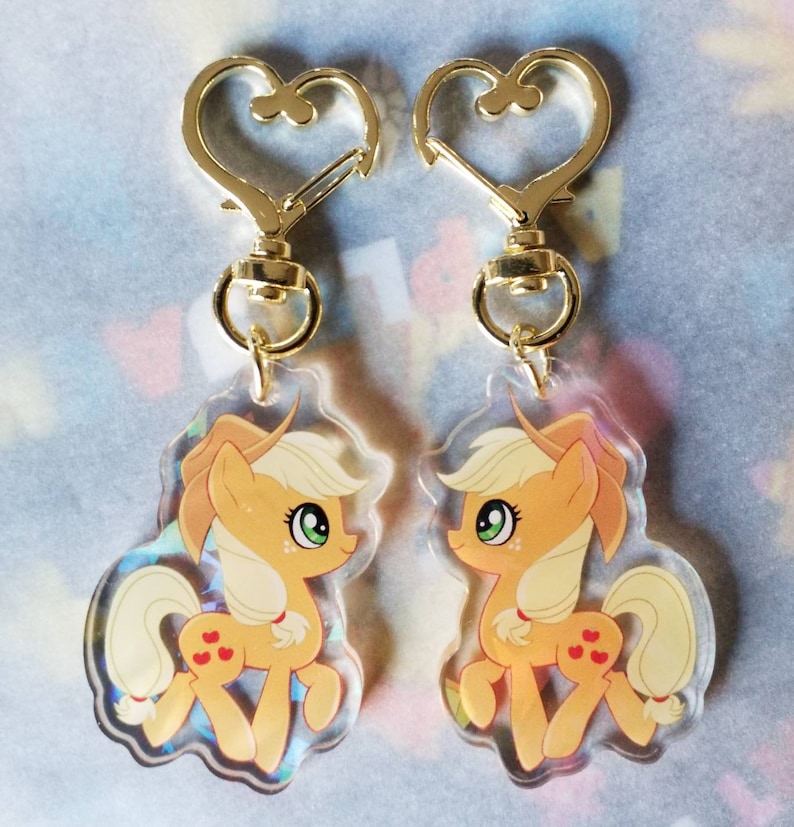 May include: Two gold heart-shaped keychains with clear acrylic charms featuring a cartoon image of Applejack from My Little Pony. The charms are translucent and show a glittery effect.